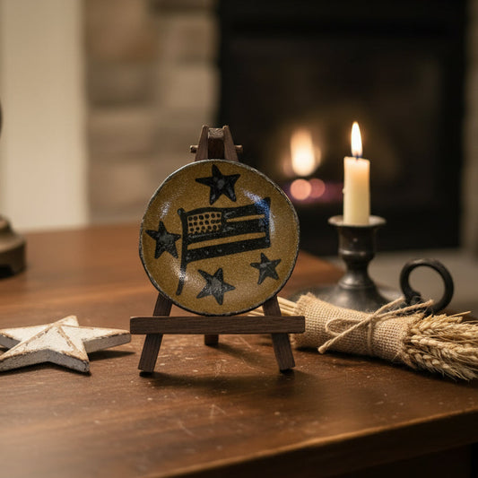A round ceramic decorative plate with a rustic hand-painted design of a bed and stars, displayed on a small wooden easel stand on a wooden table. The background features a warm, softly blurred fireplace with a lit candle and rustic decorative accents.