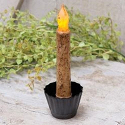 A black metal tin candle holder with a ruffled exterior, featuring a candle with a yellow flame, placed on a textured surface with greenery in the background.