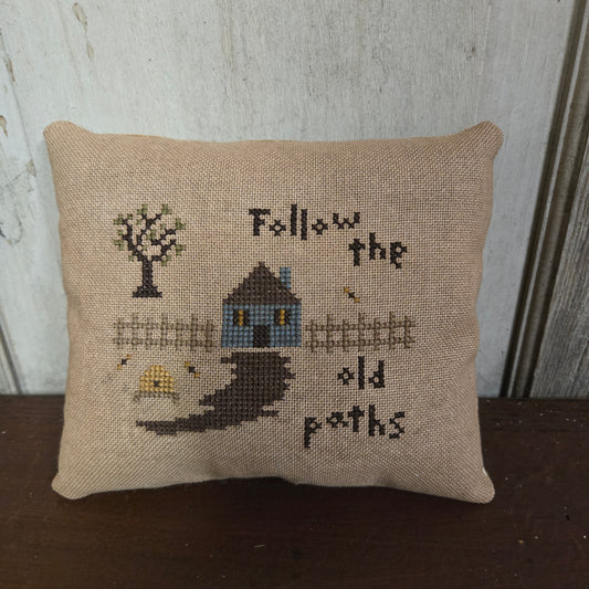 Decorative pillow with cross-stitched design and text 'Follow the old paths' on a wooden surface.