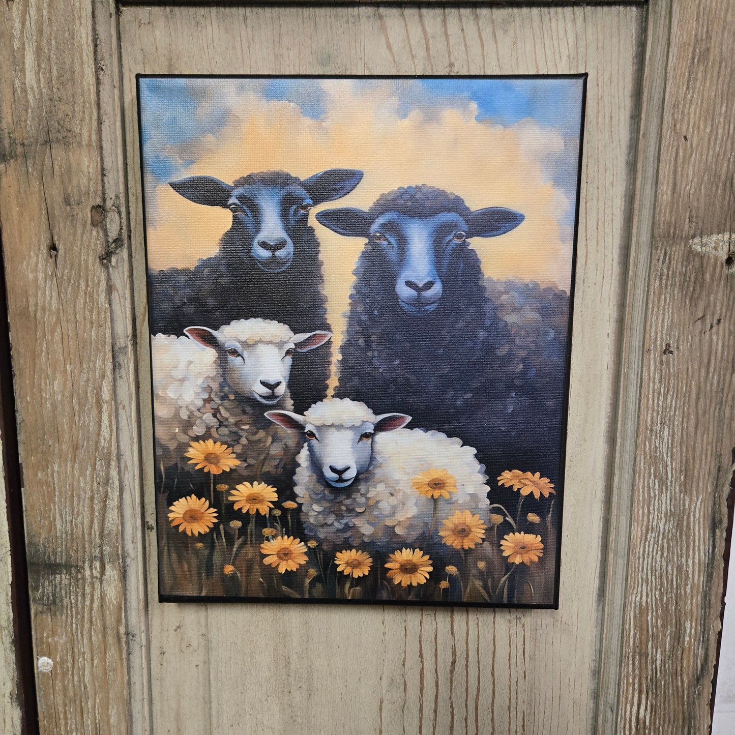 A canvas print featuring three black sheep and one white sheep among yellow flowers, set against a blue and yellow sky, displayed on a wooden surface.