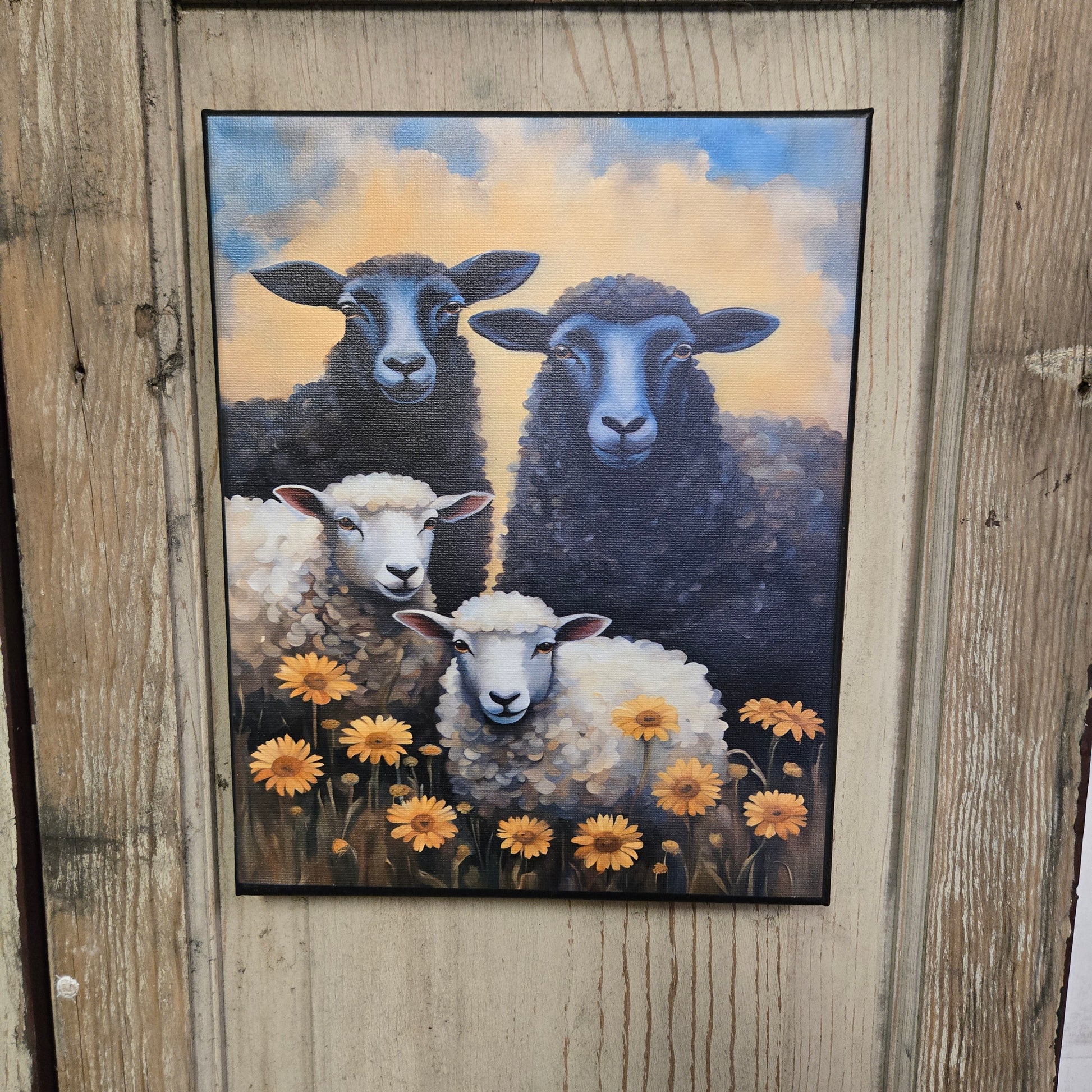A canvas print featuring three black sheep and one white sheep among yellow flowers, set against a blue and yellow sky, displayed on a wooden surface.