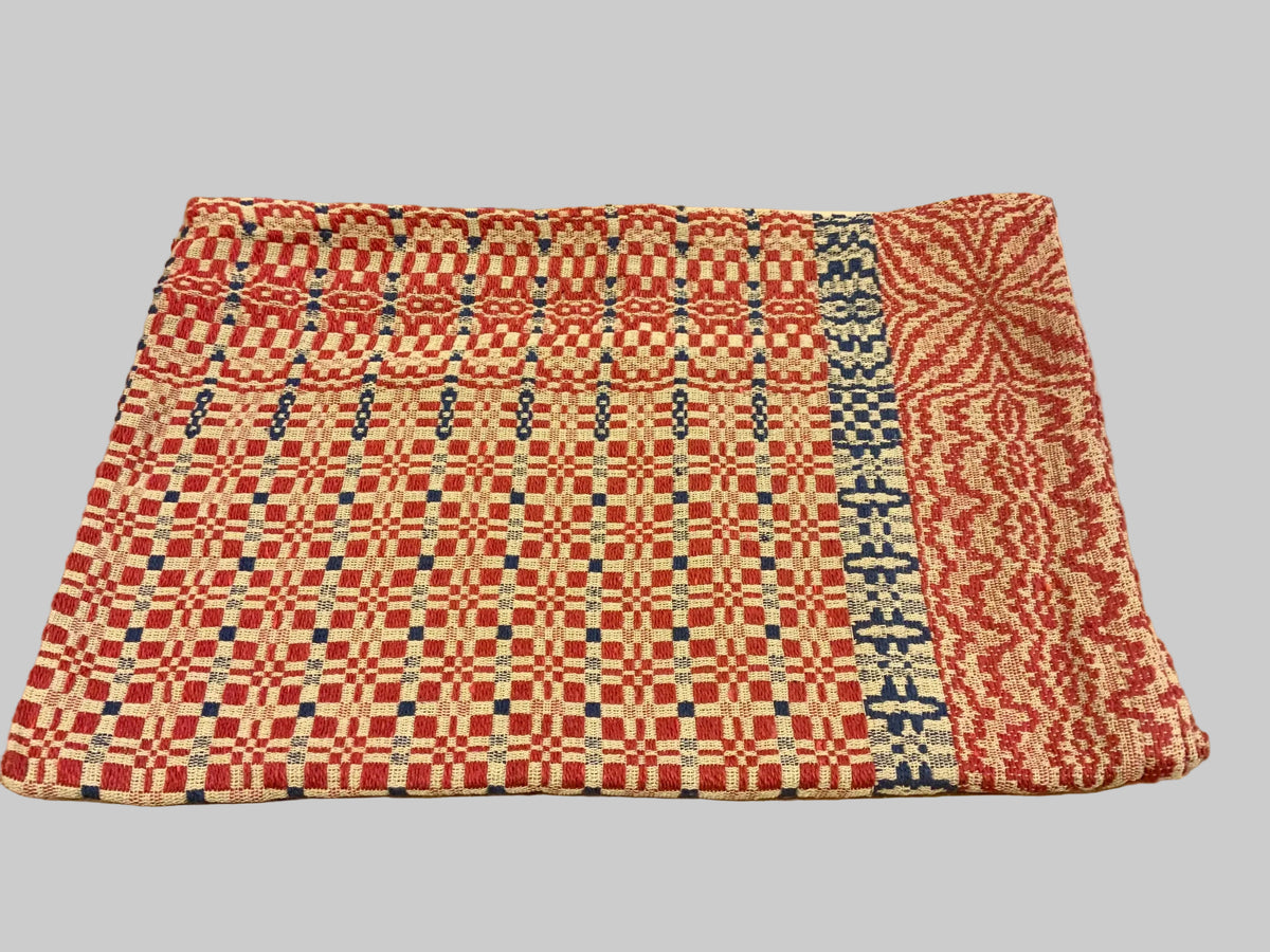 A rectangular woven textile with a red, beige, and blue geometric pattern, laid flat on a rustic wooden table in a shop setting.