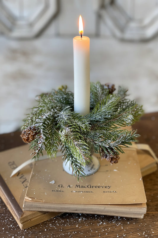 Candle in a holder made of greenery and pinecones on top of vintage books with a blurred background.