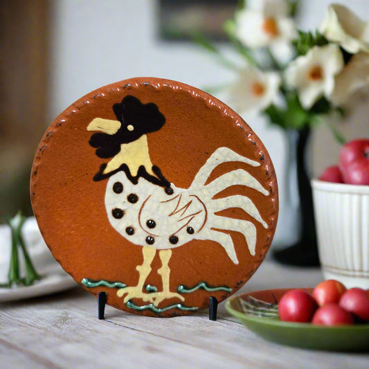 Decorative redware plate with a rooster design on a stand, set against a blurred background with flowers and apples.
