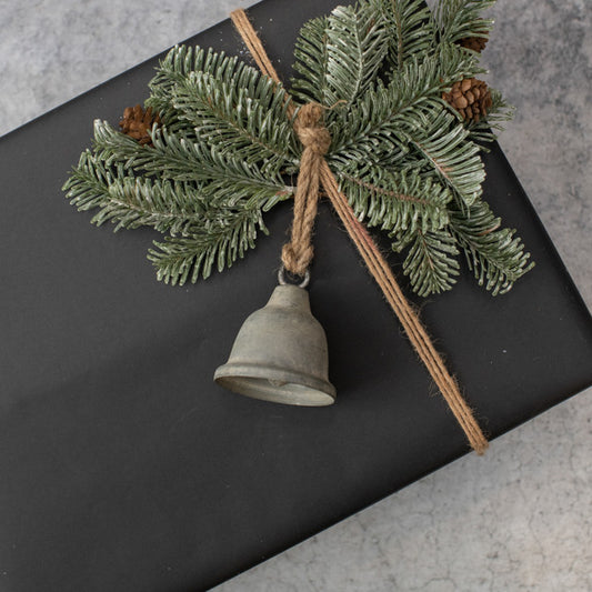 A galvanized metal bell ornament with a green pine branch and brown twine, placed on a black textured surface.