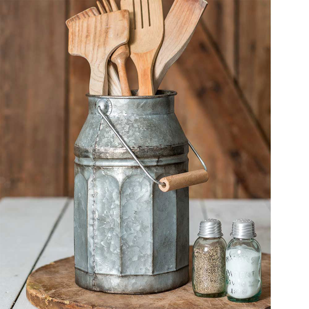 A galvanized metal milk can with a handle, containing wooden utensils, set on a wooden surface with a rustic background.