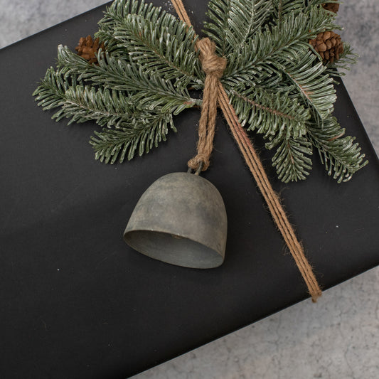 A silver galvanized Morgan bell ornament with a rustic finish, decorated with a pine branch and attached to a rope, placed on a black surface.