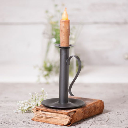 A smokey black finish candle holder with a taper candle placed in it, set on an open book, with a background of flowers and a wooden object.