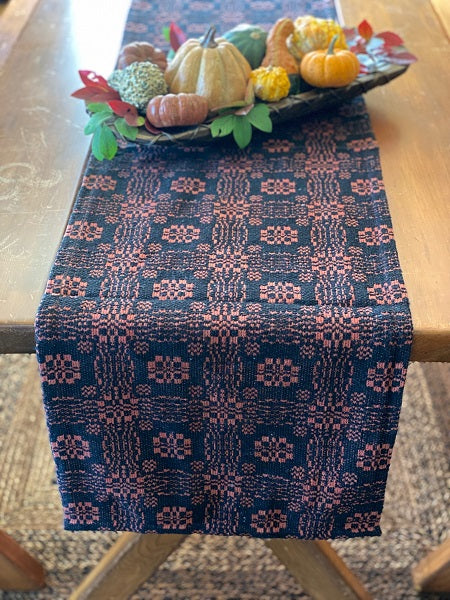 A short table runner with a geometric pattern featuring pumpkins and leaves, primarily in black and orange colors, laid out on a wooden table.