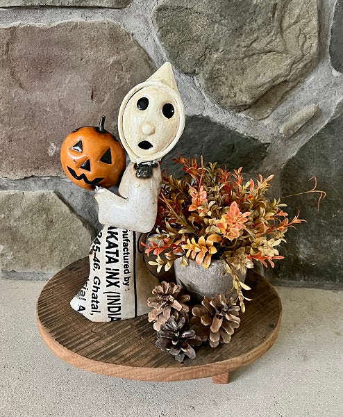 Decorative figure with a pumpkin and newspaper, placed on a wooden stand against a stone wall.