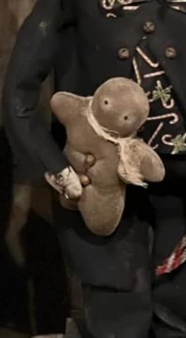 A beige stuffed gingerbread figure wearing a black coat with a floral pattern, holding a bow, and decorated with holiday-themed badges, being used as a bow filler for a bowl.