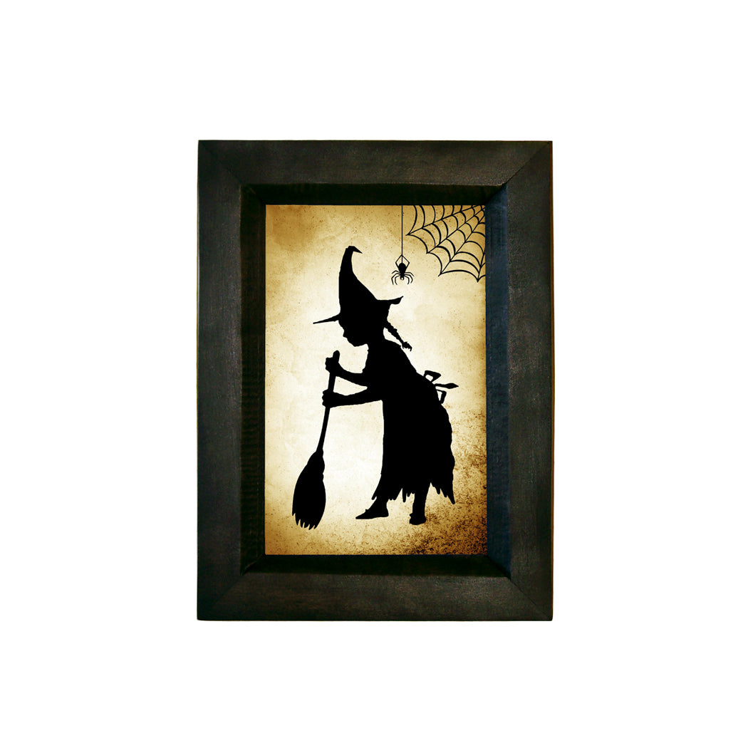 A black and white silhouette of a witch riding a broom, framed in a solid wood distressed black frame, with a spider web in the background.