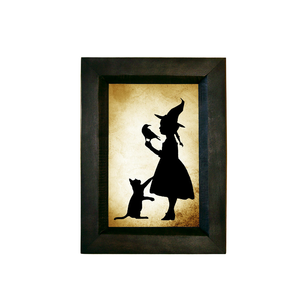A silhouette of a girl witch holding a raven on her shoulder, printed on paper, framed in a black solid wood frame.
