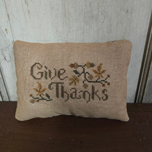 Small embroidered pillow with 'Give Thanks' text on a wooden surface