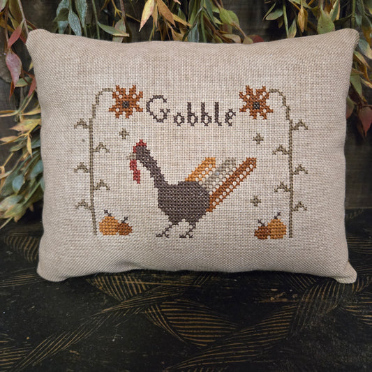 Decorative pillow with cross-stitched design of a turkey and 'Gobble' text, surrounded by autumn leaves.