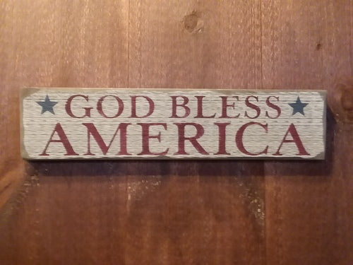 A wooden sign with 'GOD BLESS AMERICA' text in white and red on a blue background, mounted on a wall.