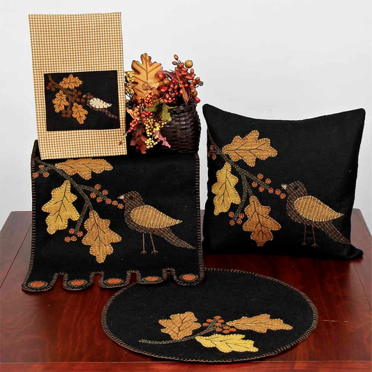 A set of table runners and pillows with a bird and leaf design, primarily in black, mustard, and golden colors, displayed on a wooden table.