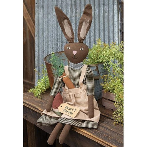 A decorative plush bunny made of brown linen fabric with a painted face, dressed in a black and white gingham dress with a taupe apron, holding a bundle of fruits and vegetables, sitting against a rustic background.