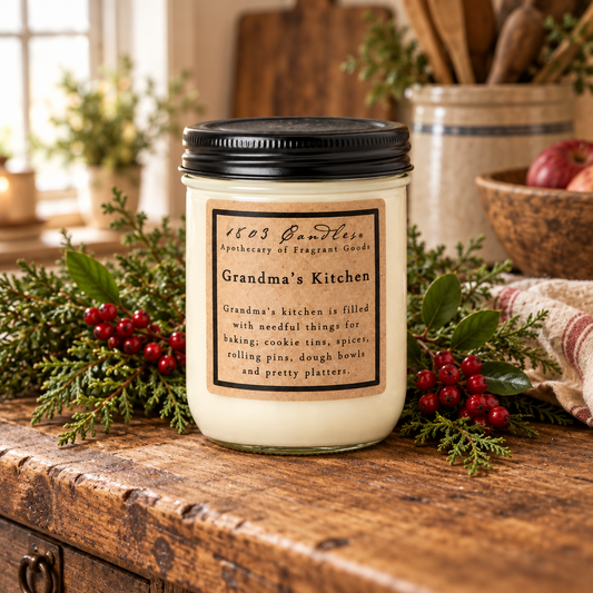A glass jar candle with a black lid and a tan label reading '1803 Samtner Anniversary of Scenic Candle Grandma's Kitchen' sits on a rustic wooden table surrounded by greenery and red berries in a cozy kitchen setting.