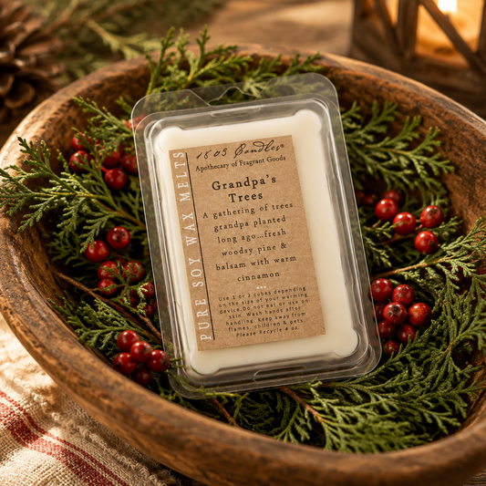 A clear plastic clamshell tub of wax melts sits in a rustic wooden bowl surrounded by evergreen branches and red berries. The label reads '1623 of Lee Apothecary of Tregellis' and 'Grandpa's Trees' with a scent description mentioning cinnamon and warm pine tones.