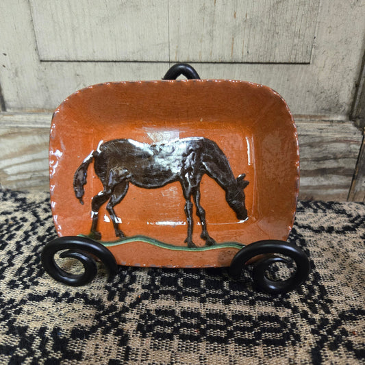 A rectangular ceramic plate with a brown horse, displayed on a wooden surface with a textured black and white background.