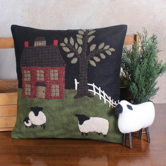 Wool felt pillow with a countryside scene including a red house, tree, and sheep in green and black colors.