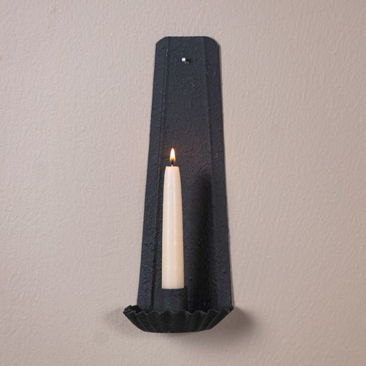 A black, traditional candle sconce mounted on a wall with a candle lit and glowing.