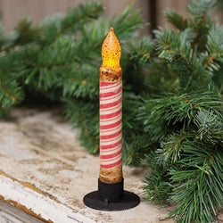 A tapered candle with candy cane stripes and a glittery, rustic finish, placed on a surface with pine branches and needles in the background.