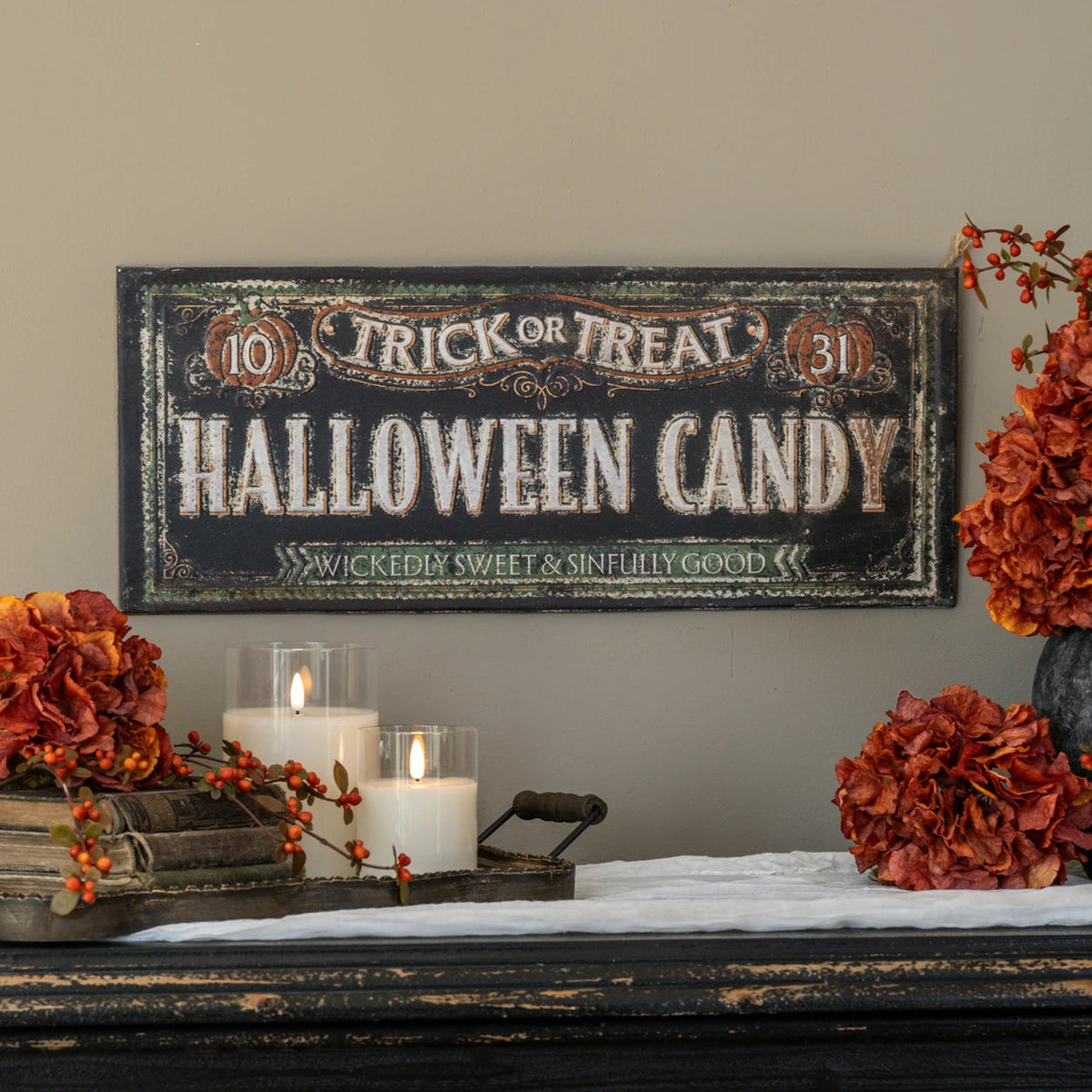 A rustic black wooden sign reading 'Trick or Treat Halloween Candy' with distressed white lettering and orange decorative accents, displayed on a mantel with candles, flowers, and fall decor against a beige wall.