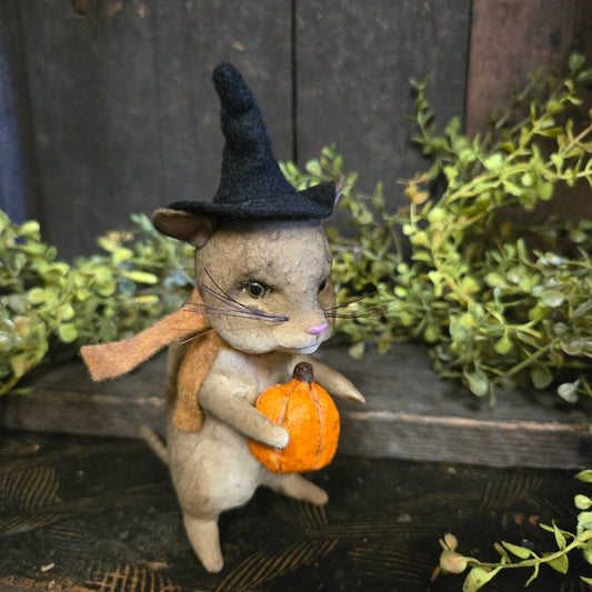 Stuffed animal resembling a mouse holding an orange pumpkin with greenery in the background