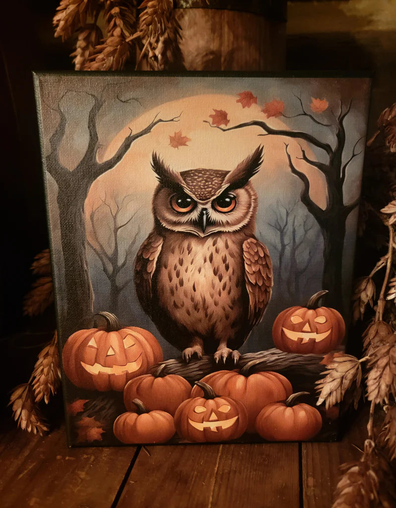 A square canvas painting of an owl perched on jack-o'-lantern pumpkins in a spooky forest scene with bare trees and a full moon, displayed on a wooden surface with dried autumn decor around it.