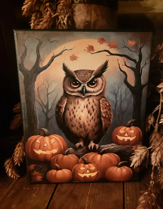 A square canvas painting of an owl perched on jack-o'-lantern pumpkins in a spooky forest scene with bare trees and a full moon, displayed on a wooden surface with dried autumn decor around it.