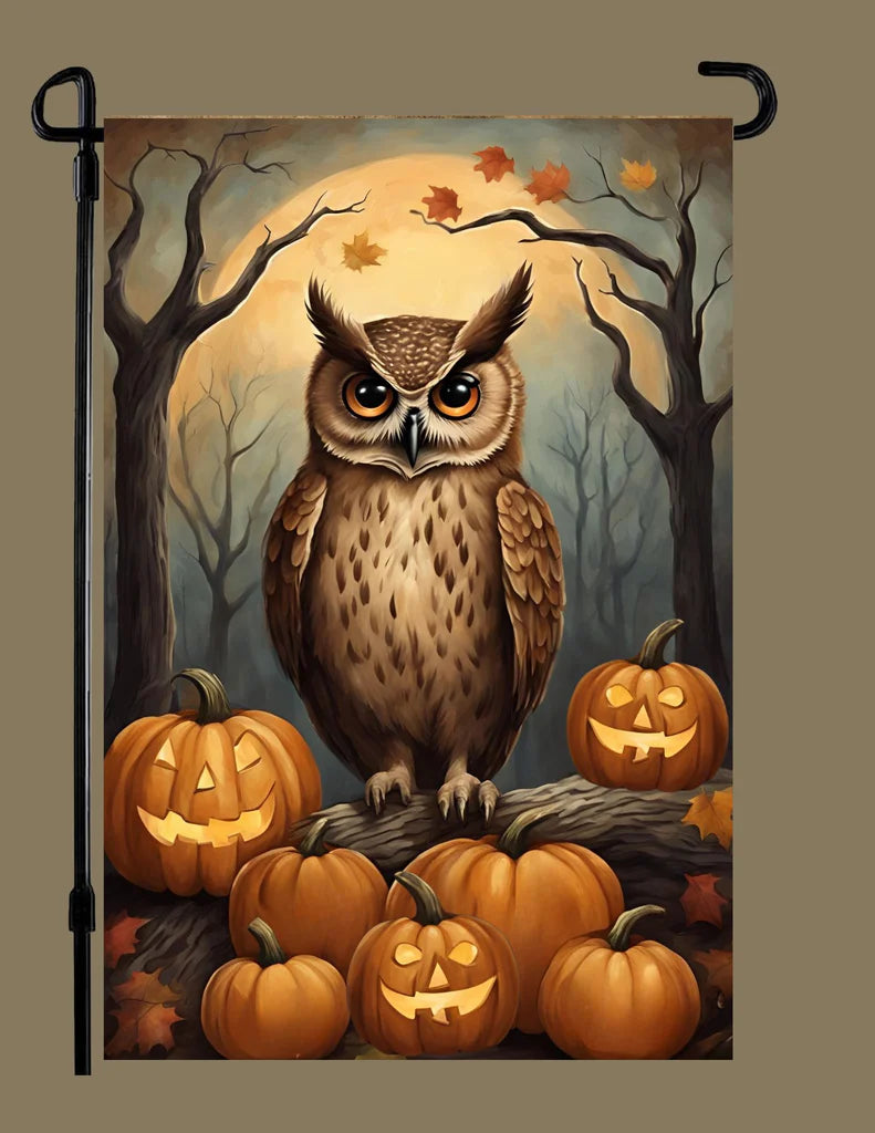 A vertical garden flag featuring a spooky Halloween illustration of a large owl perched on jack-o'-lantern pumpkins in a dark forest with bare trees and a glowing full moon. The flag is shown hanging from a black metal stand against a solid taupe background.