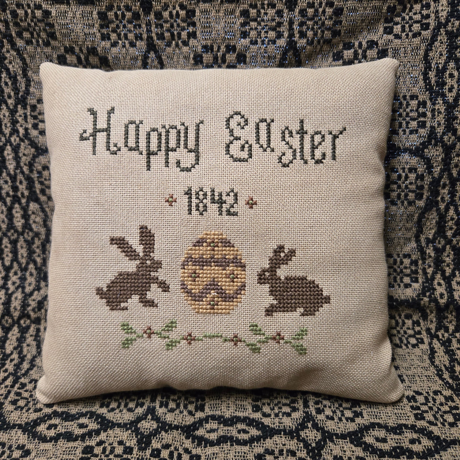 Decorative pillow with cross-stitched design of a rabbit and text 'Happy Easter 1842' on a textured fabric background.
