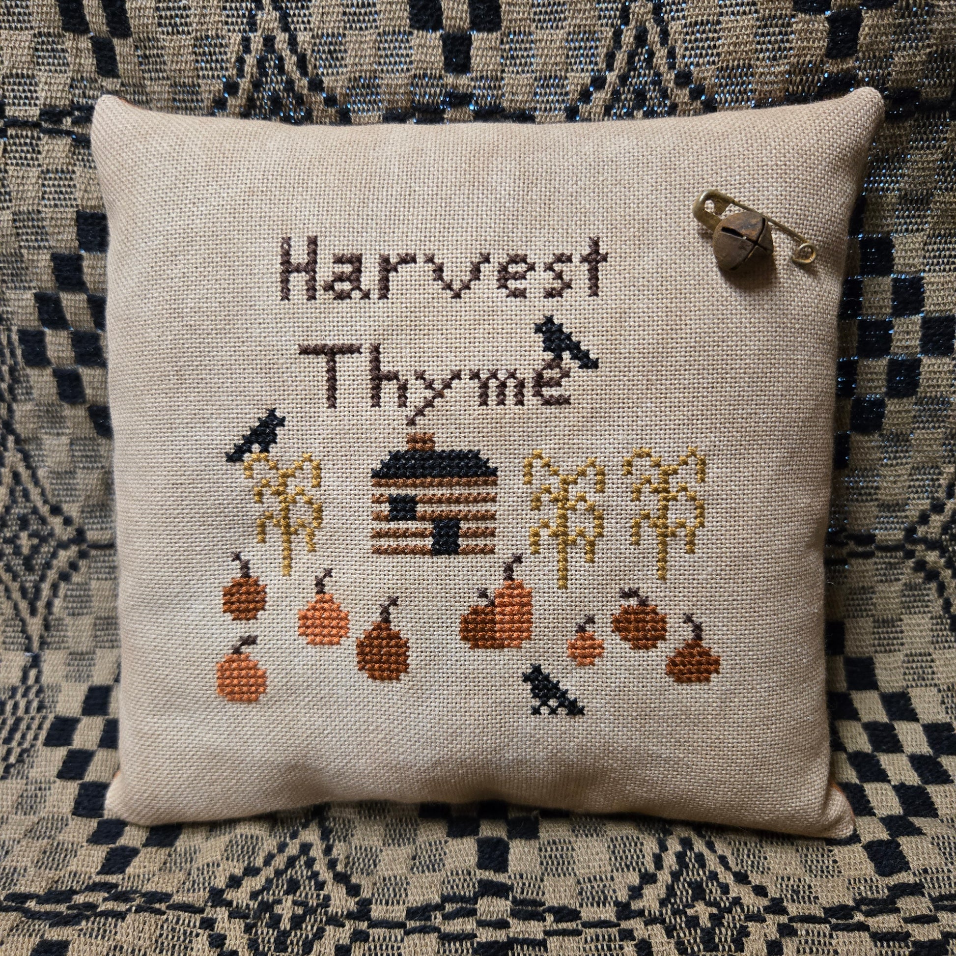 A square beige cross-stitch pillow featuring the words 'Harvest Thyme' and an autumn-themed design with pumpkins, crows, and a small house. The pillow is displayed on a dark patterned woven blanket with a silver safety pin attached near the top right corner.