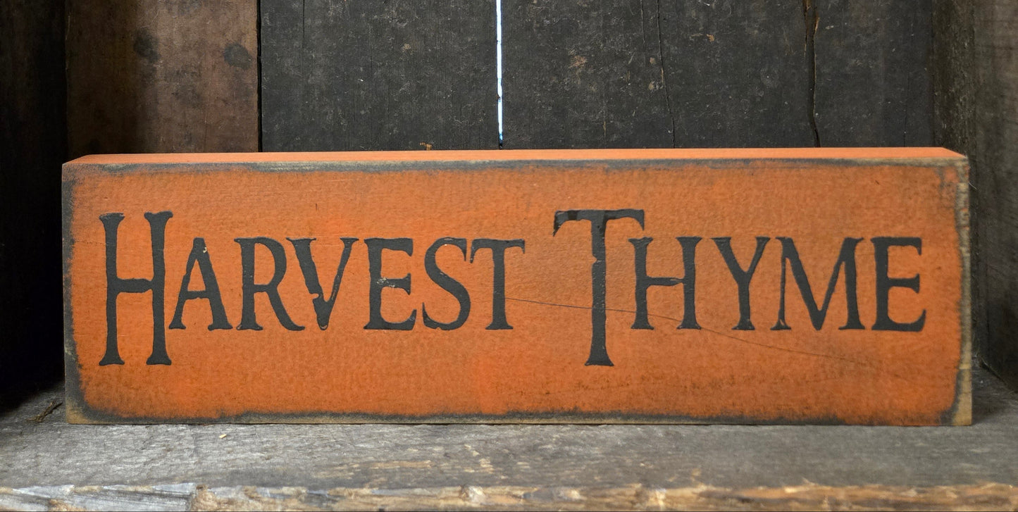 A rustic rectangular wooden sign painted orange-brown with the words “HARVEST THYME” in dark lettering, placed against a dark weathered wood background.