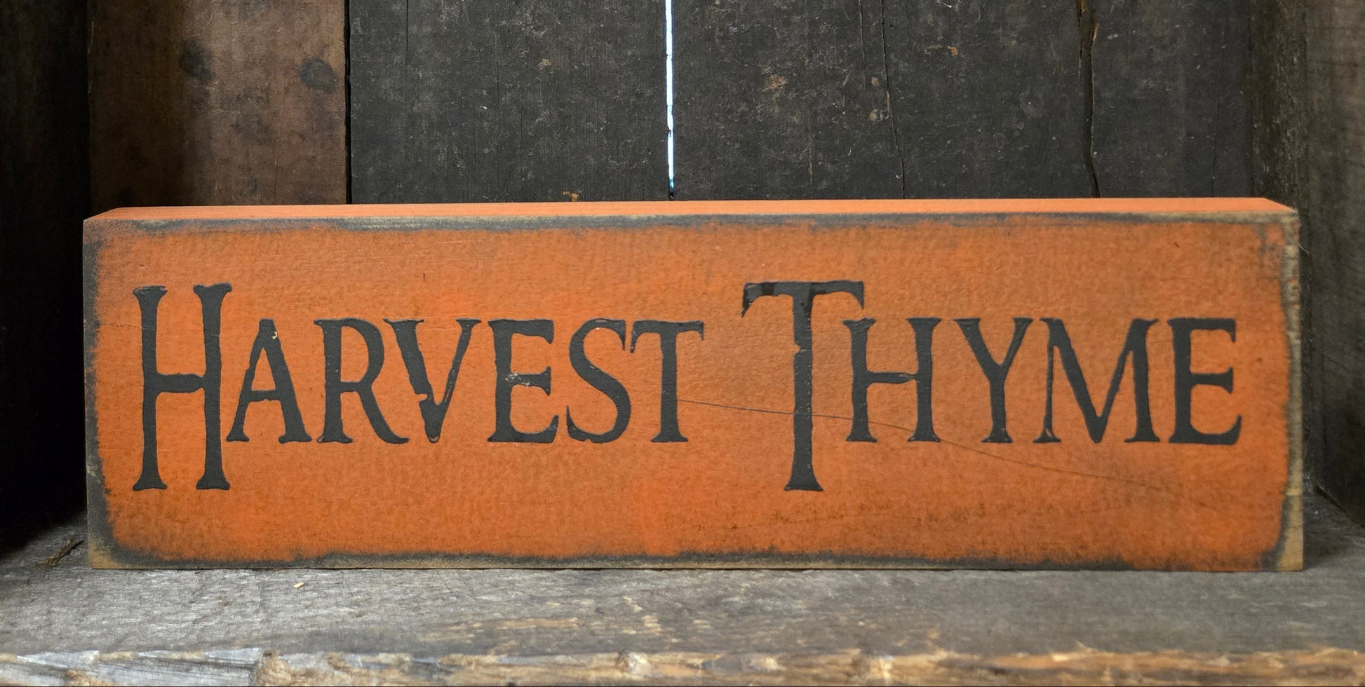 A rustic rectangular wooden sign painted orange-brown with the words “HARVEST THYME” in dark lettering, placed against a dark weathered wood background.
