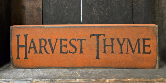A rustic rectangular wooden sign painted orange-brown with the words “HARVEST THYME” in dark lettering, placed against a dark weathered wood background.