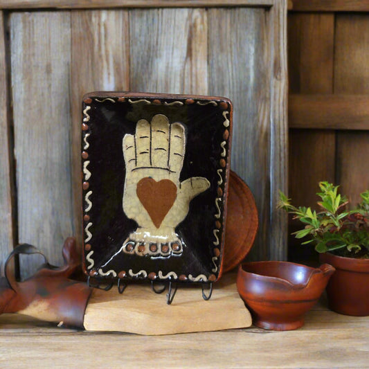 Redware plate with a hand and heart design on a wooden stand, surrounded by rustic decor.