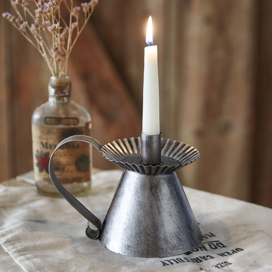 A metal chamberstick candle holder with a lit candle, placed on a table with a vintage bottle and flowers in the background.
