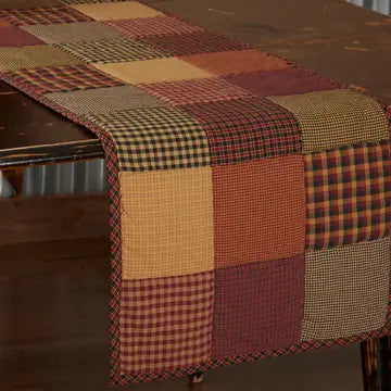 A rustic patchwork table runner draped over a dark wooden table, featuring a mix of gingham, plaid, and solid fabric squares in autumnal colors like rust, terracotta, mustard, and brown.