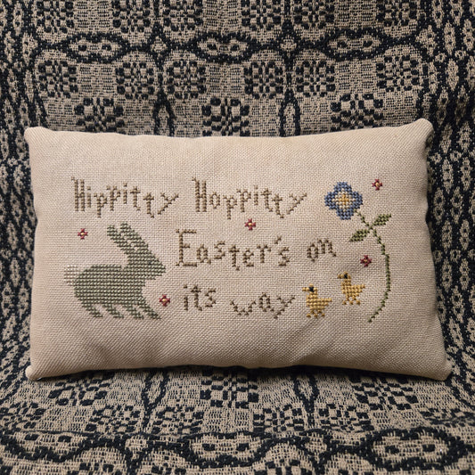 Decorative pillow with Easter-themed cross-stitch text and illustrations on a patterned fabric background.