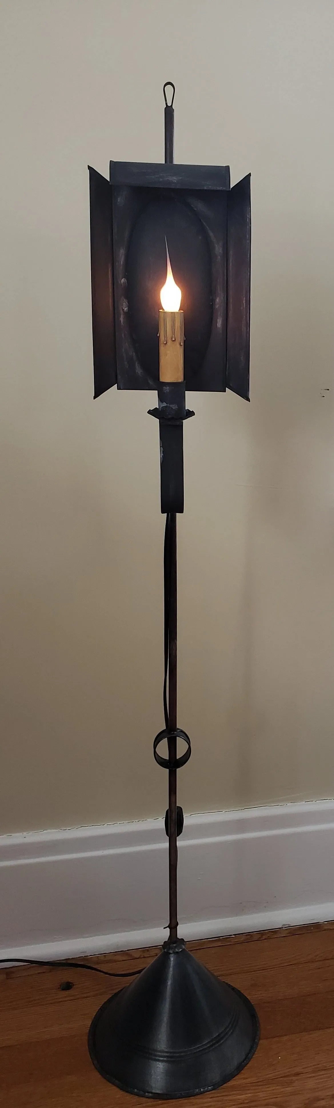 A Hobart Mini Tin Floor Lamp with a black primitive tin finish, featuring a candle-like light source, mounted on a circular black base.