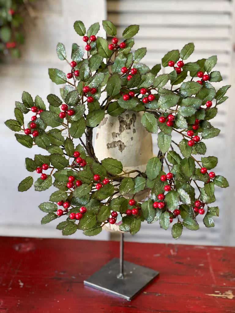 Decorative wreath with green leaves and red berries on a stand against a neutral background.