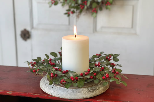 Decorative candle holder with a white candle, surrounded by greenery and red berries, on a wooden surface.
