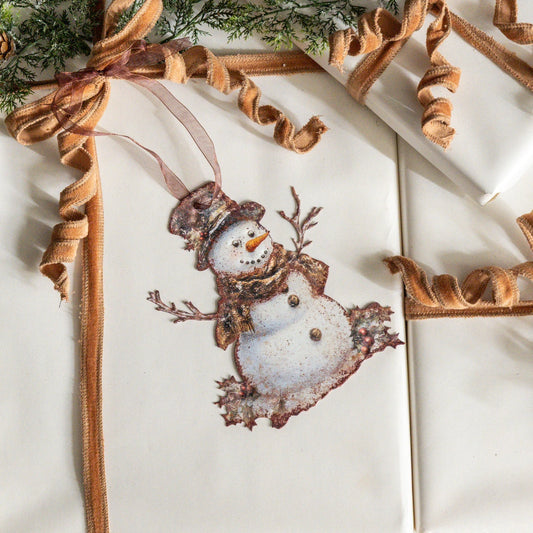 A close-up of a wrapped gift box or card featuring a printed snowman ornament design in the center, tied with a tan ribbon and surrounded by similar wrapped packages and ribbon.