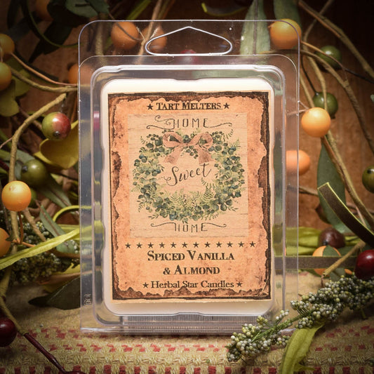 A clear plastic clamshell packet containing a mini wax tart with a label that reads 'Spiced Vanilla & Almond - Herbal Star Candles'. The packet is displayed in front of a decorative background with fruit and foliage.