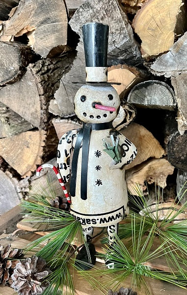 Decorative snowman figurine with a top hat and scarf, standing on wood and pine branches.