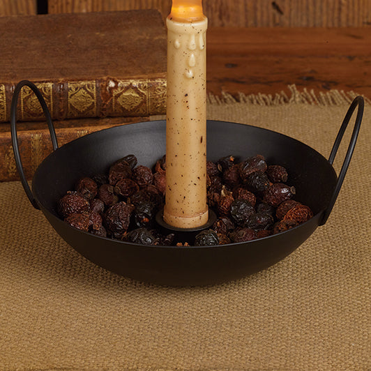 A black metal tapered baking pan with two handles, filled with roasted nuts and a candle placed in the center, set against a backdrop of books.