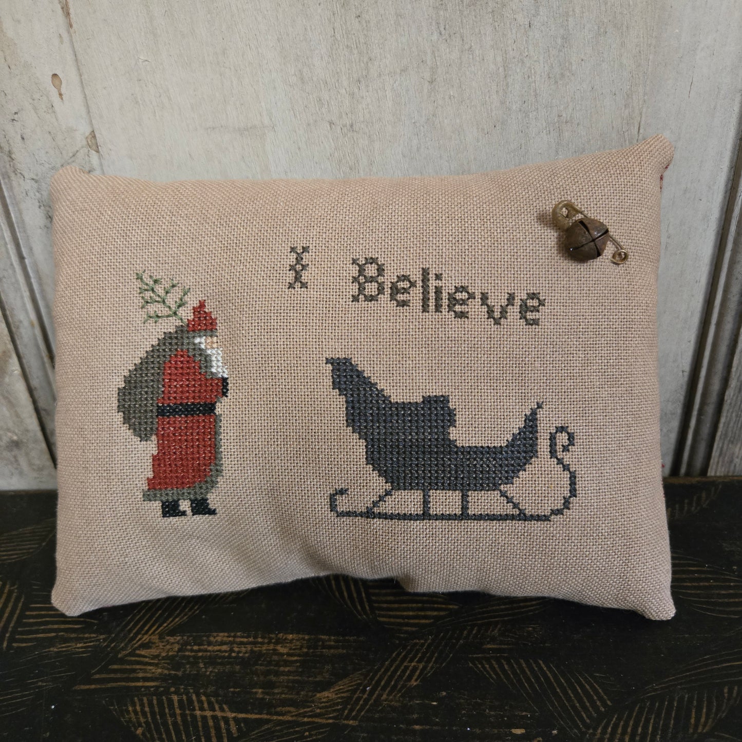 Decorative pillow with cross-stitched design of a sleigh, Santa Claus, and the word 'Believe' on a wooden surface.